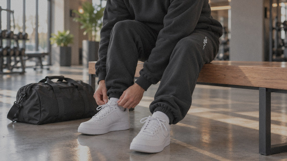 Person in a black heavyweight hoodie and joggers tying white sneakers on a gym bench next to a duffel bag