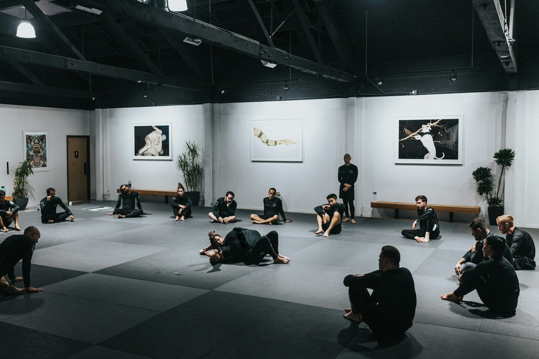 No-gi BJJ class sitting around the mats while two students demonstrate a technique in a bright academy