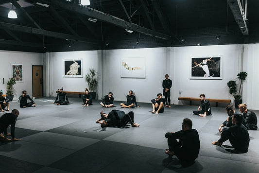 No-gi BJJ class sitting around the mats while two students demonstrate a technique in a bright academy