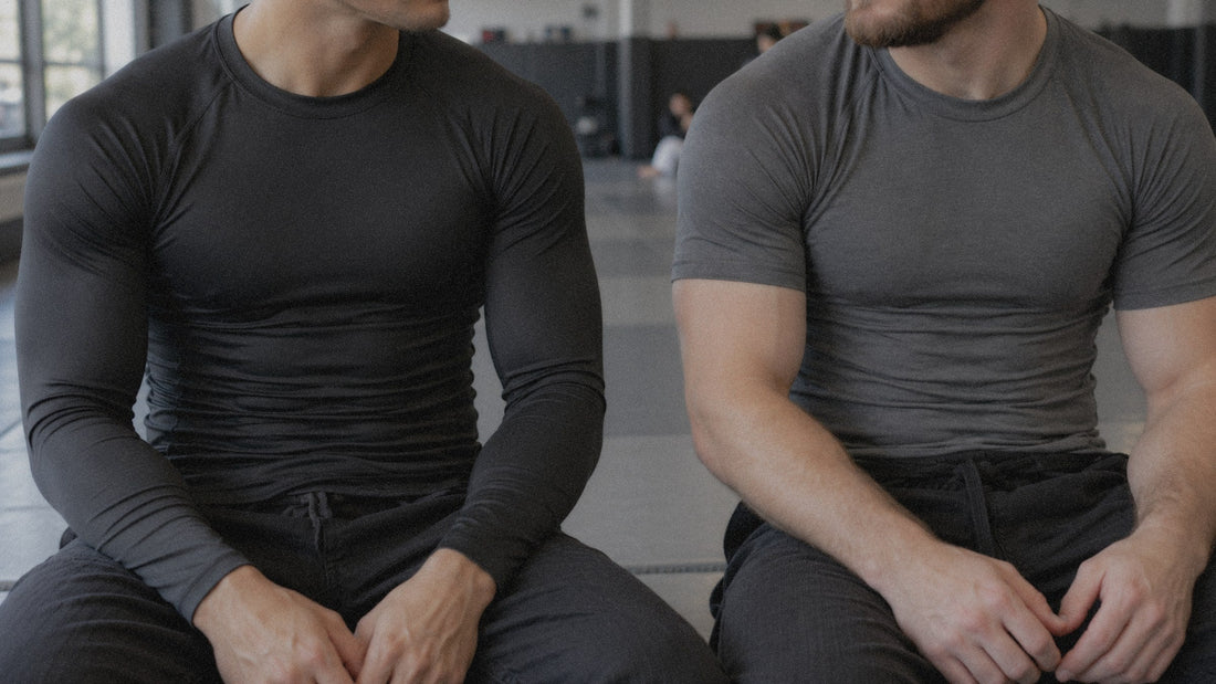 Two grapplers seated on the mat, one in a black long sleeve rashguard and one in a gray compression shirt